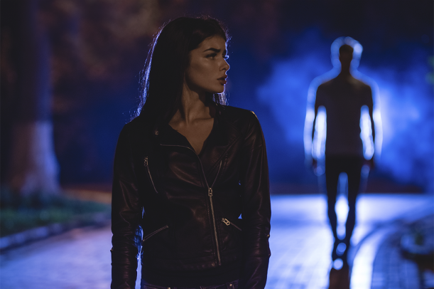 A woman walking alone at night on a lit path with another figure approaching in the distance behind her