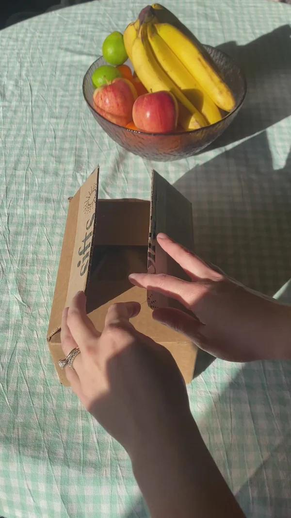 Hands opening a branded Sifts box with fruit in the background
