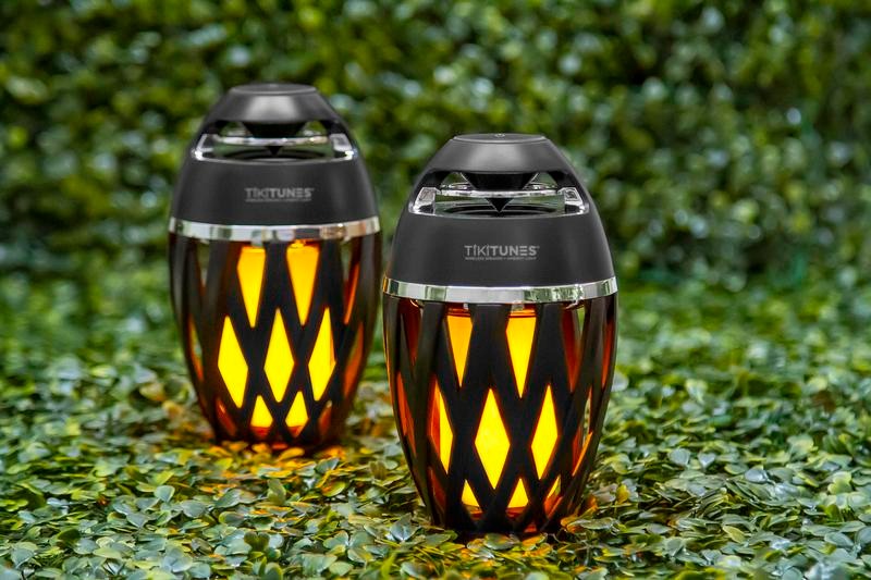 Two TikiTunes wireless speakers with ambient flame lights placed on green foliage