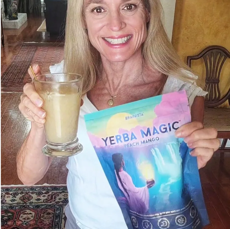 A smiling woman holding a glass of prepared drink and a bag of Yerba Magic Peach Mango